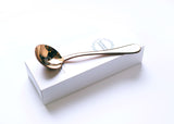 A copper colored coffee tasting spoon on top of its white box. The spoon says Fair For All on the end of the handle and has a round Olympia Coffee logo in the bowl of the spoon. The white box fits the spoon and has the Olympia Coffee logo near the top edge and says "Coffee Cupping Spoon" near the bottom edge.