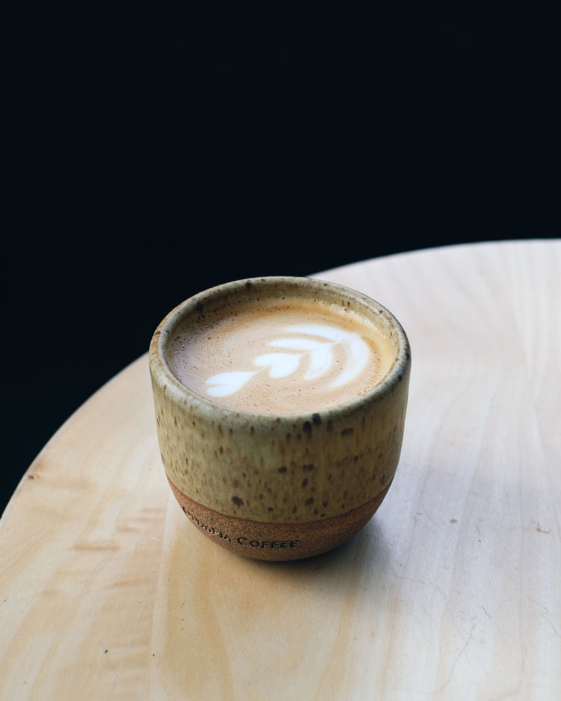 One 6 oz. cappuccino cup by Olympia-based potter Baker|Potter. The cup is filled with a cappuccino, with flower latte art in the foam. The cup is sitting on a cafe table.
