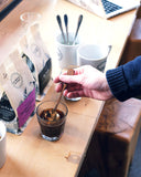 A barista dipping the coffee tasting spoon into a glass of brewed coffee. Two bags of coffee, and three Olympia Coffee logo mugs sit nearby, one of which has three spoons sitting in it.