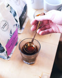 Close up of a barista dipping the coffee tasting spoon into a glass of brewed coffee. Two bags of coffee and two Olympia Coffee logo mugs sit nearby on the counter.