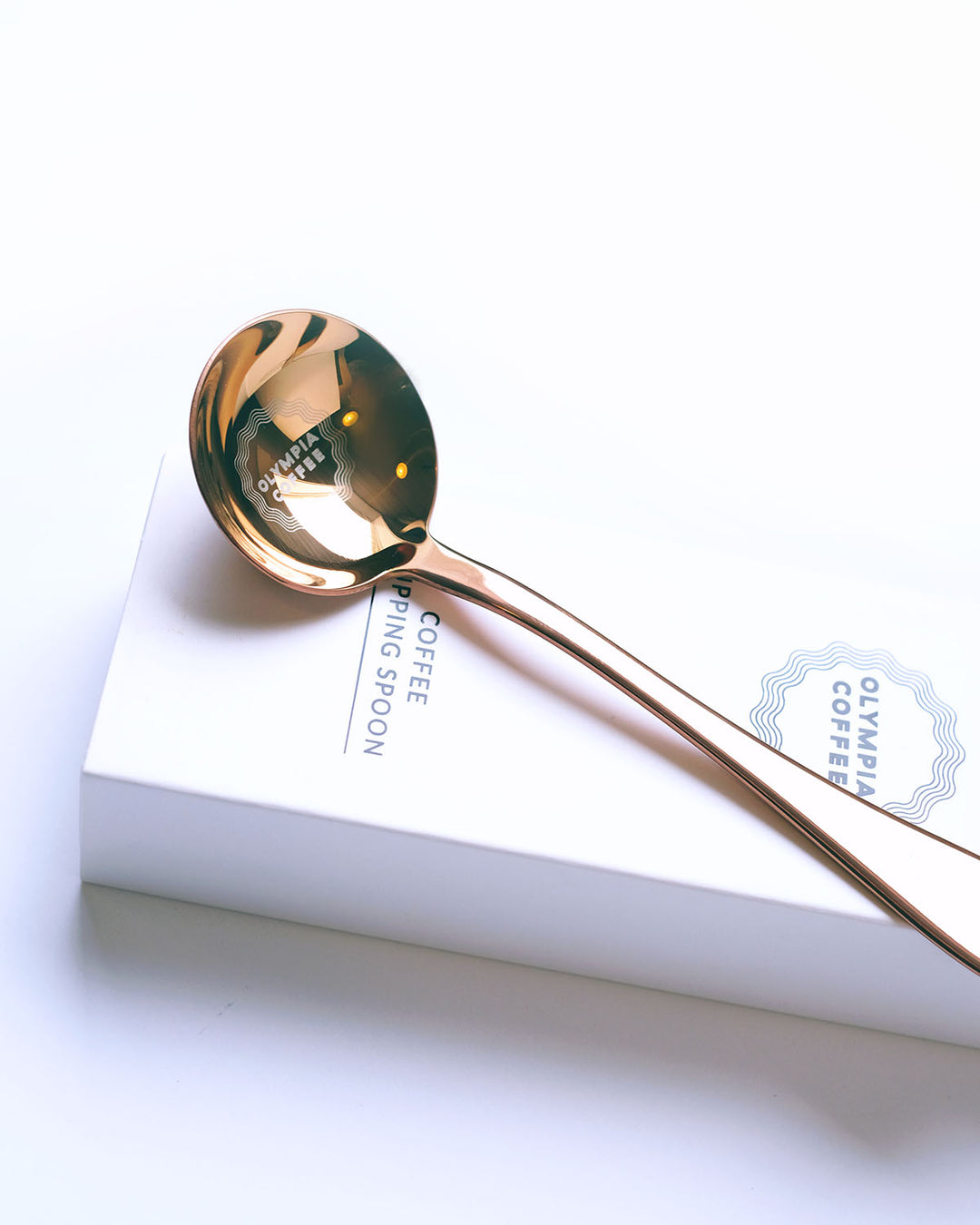 A copper colored coffee tasting spoon on top of its white box. The spoon says Fair For All on the end of the handle and has a round Olympia Coffee logo in the bowl of the spoon. The white box fits the spoon and has the Olympia Coffee logo near the top edge and says "Coffee Cupping Spoon" near the bottom edge.