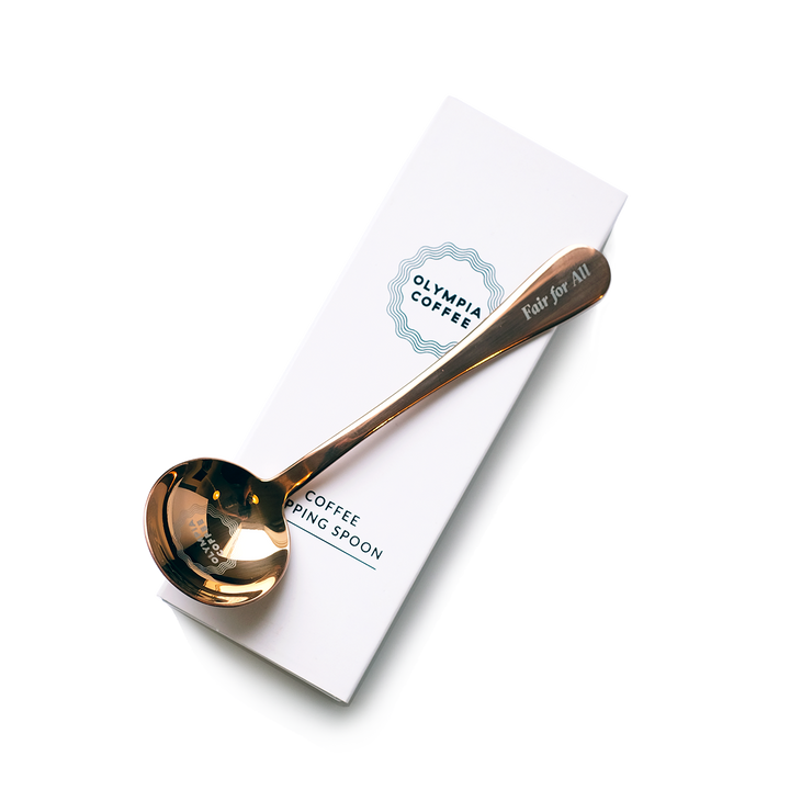 A copper colored coffee tasting spoon on top of its white box. The spoon says Fair For All on the end of the handle and has a round Olympia Coffee logo in the bowl of the spoon. The white box fits the spoon and has the Olympia Coffee logo near the top edge and says "Coffee Cupping Spoon" near the bottom edge.