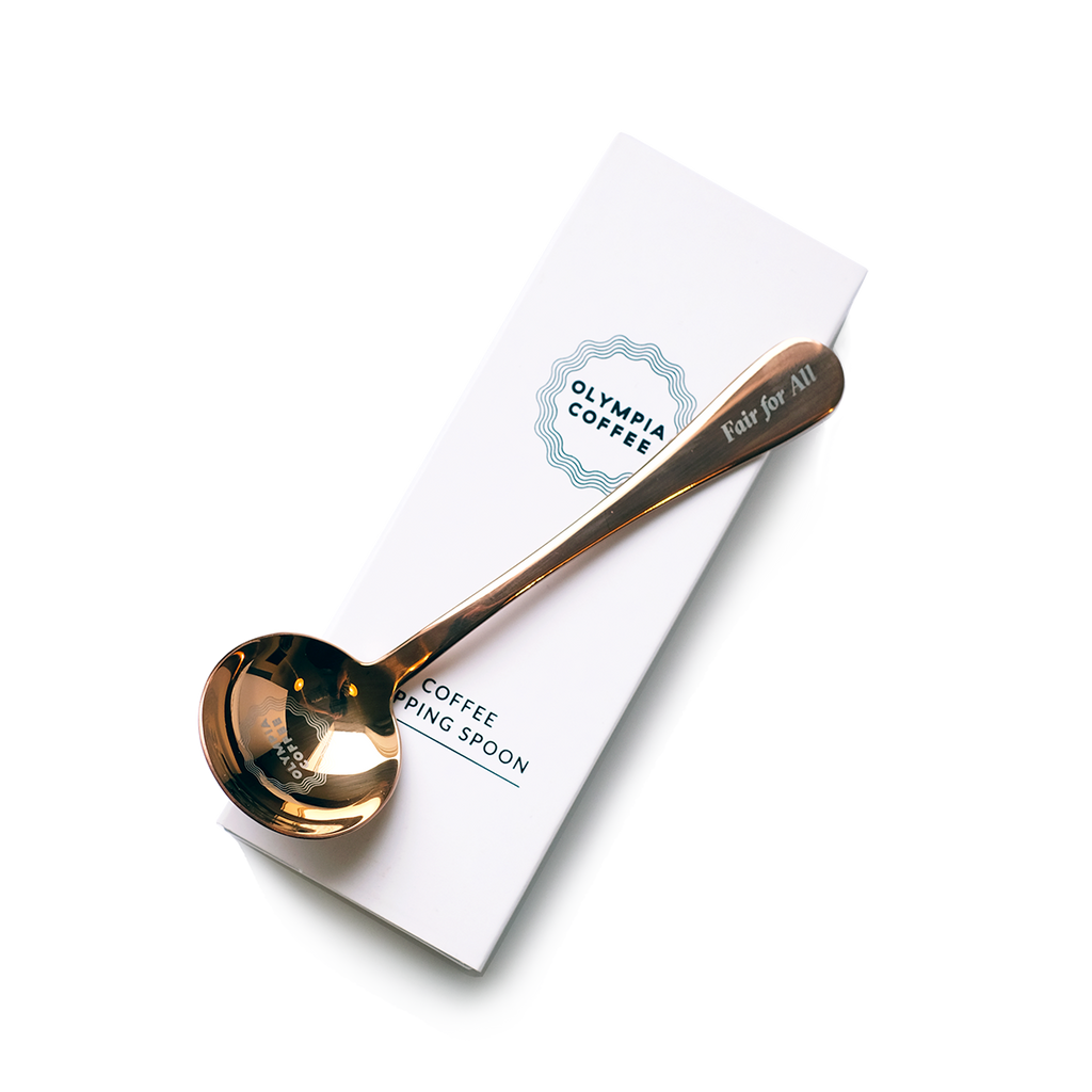 A copper colored coffee tasting spoon on top of its white box. The spoon says Fair For All on the end of the handle and has a round Olympia Coffee logo in the bowl of the spoon. The white box fits the spoon and has the Olympia Coffee logo near the top edge and says "Coffee Cupping Spoon" near the bottom edge.