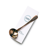A copper colored coffee tasting spoon on top of its white box. The spoon says Fair For All on the end of the handle and has a round Olympia Coffee logo in the bowl of the spoon. The white box fits the spoon and has the Olympia Coffee logo near the top edge and says "Coffee Cupping Spoon" near the bottom edge.